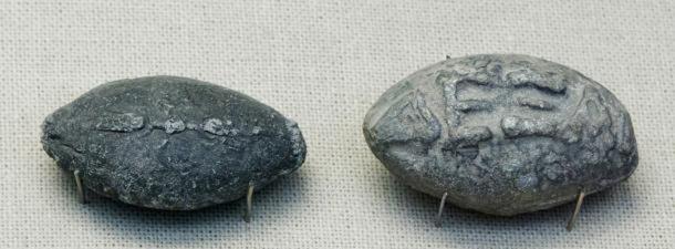 Ancient Greek sling bullets with engravings. One side depicts a winged thunderbolt, and the other, the Greek inscription “take that” in high relief.