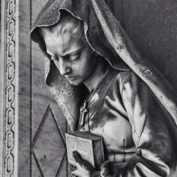 A woman holds a bible close to her chest as she lowers her head in grief (lnm / Adobe Stock)
